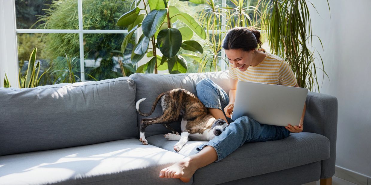 Woman laughing with her dog on a couch with a laptop.