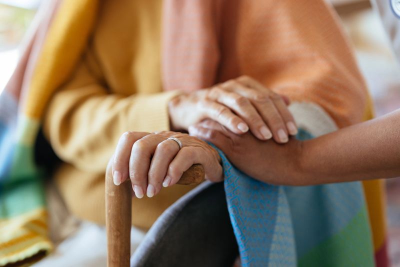 A close-up of an elderly person's hand resting on a wooden cane, with a caregiver’s hand gently placed on top in a comforting gesture. The senior is wrapped in a colorful blanket, symbolizing warmth, care, and support in a home or healthcare setting