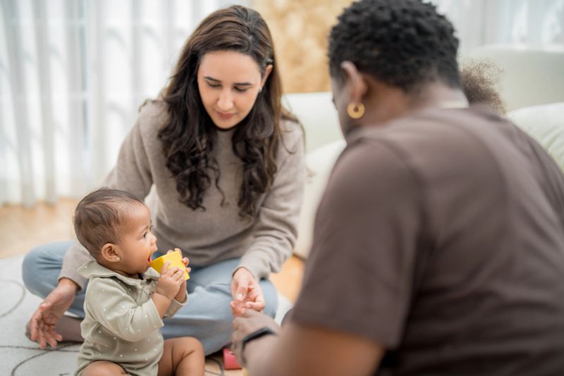 A toddler plays with a bright yellow cup while sitting on a soft rug. Two adults engage with joy, encouraging the child's exploration and interaction in a warm, inviting atmosphere.