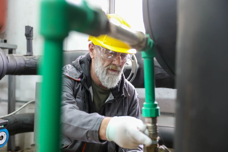 A senior male engineer with a gray beard, wearing a yellow hard hat, safety goggles, white protective gloves, and a dark work jacket, is adjusting the settings on an industrial heat pump control panel. He is holding a tablet in one hand while pressing a red emergency stop button with the other. The background features an industrial setting with insulated black pipes, valves, and a fire extinguisher, indicating a technical environment focused on heating and cooling systems.