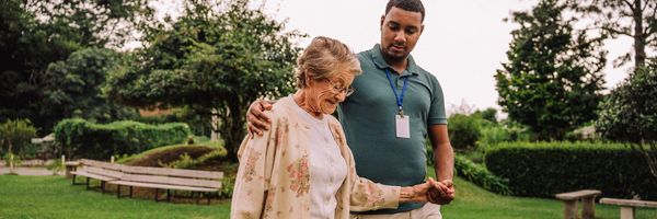 Young man supports elderly woman walking outdoors in a park.