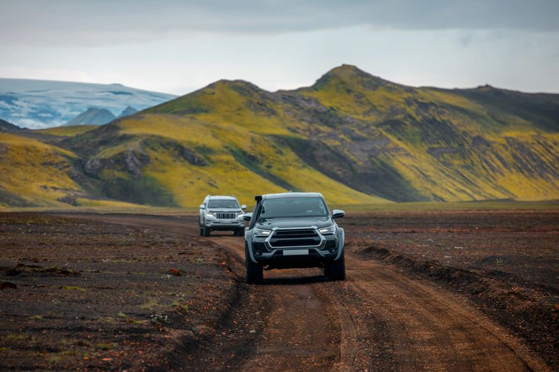 Two off-road vehicles driving along a rugged dirt road through the volcanic terrain of Iceland. The lush green moss covering the mountains contrasts sharply against the dark soil and rocky path. The scene reflects the adventurous spirit of Icelandic exploration, with glaciers visible in the background