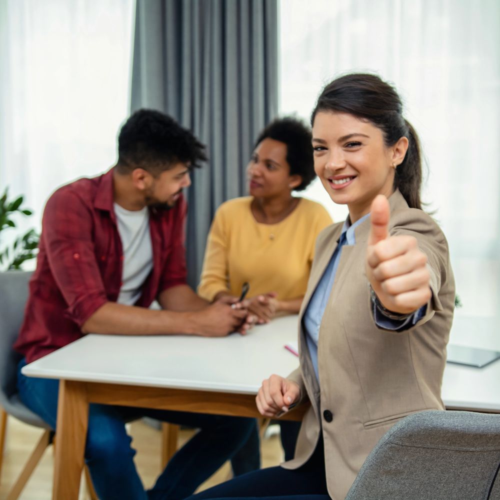 Confident woman giving a thumbs-up in a meeting with two colleagues.