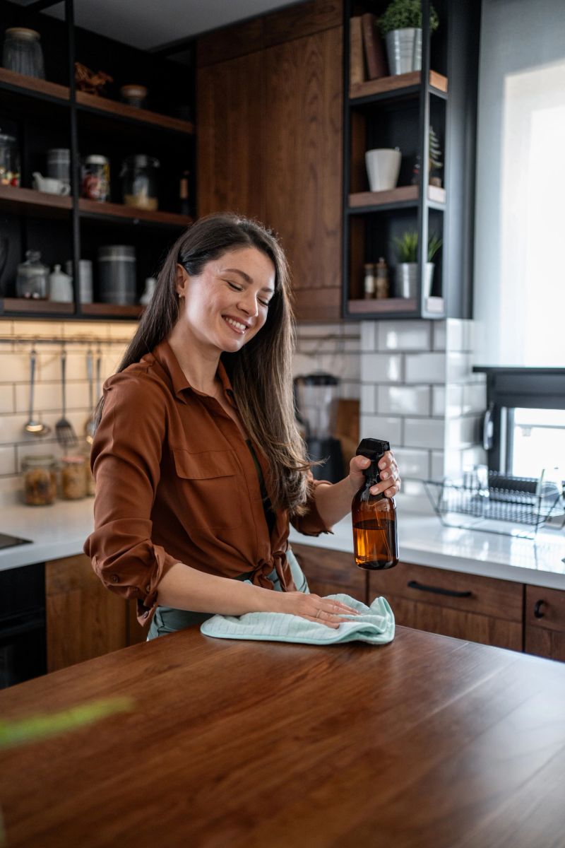 Smiling young woman spraying a wooden kitchen counter with an eco-friendly cleaning product while wiping it down with a cloth, enjoying her clean and organized home