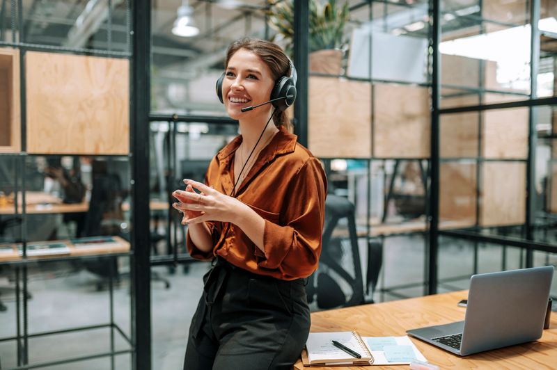 Female customer support officer, wearing headset and standing at desk, smiling cheerfully with hands clasped, listening to client online in call center