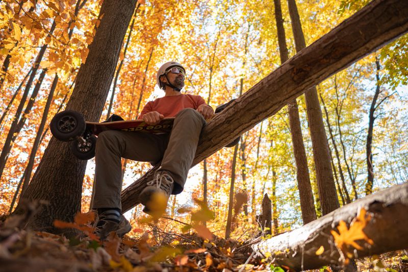 Man wearing sports helmet & sunglasses, & his electric mountain board, sits contemplatively on fallen tree trunk among vibrant autumn forest at sunset. Digital detox & peace in nature.