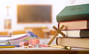 Stack of books with a wooden cross and school supplies on a desk.
