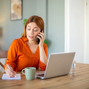 Woman in orange shirt talking on phone and taking notes at a wooden table.