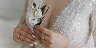 Bride holding a white floral boutonniere, wearing a beaded wedding dress and rings.