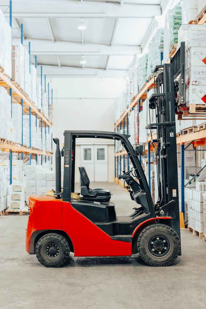 A bright red forklift is positioned centrally in a spacious industrial storage area with high shelves on both sides, loaded with pallets and packaged items. The clean and well-lit environment highlights efficiency and order within the facility. This image illustrates essential warehouse equipment, inventory handling, and the role of material transport vehicles in logistics and supply chain operations.
