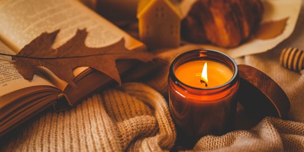 A lit candle on a cozy knitted fabric with an open book and autumn leaf.