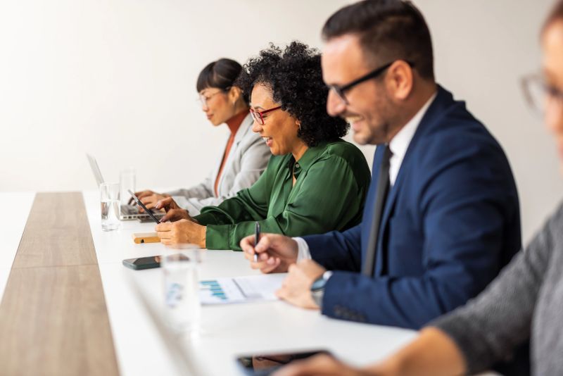 A group of corporate professionals working together at a table, conducting collaborative activities including writing, observing charts, and using digital devices. Each person is focused and engaged, fostering a productive atmosphere.