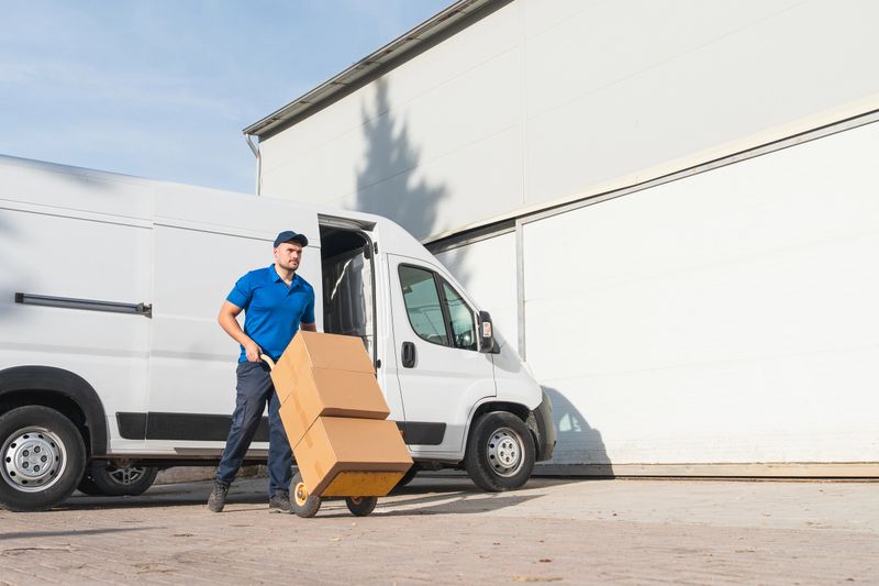 Delivery man in blue uniform transporting cardboard boxes on a hand truck outside a warehouse with a white van.