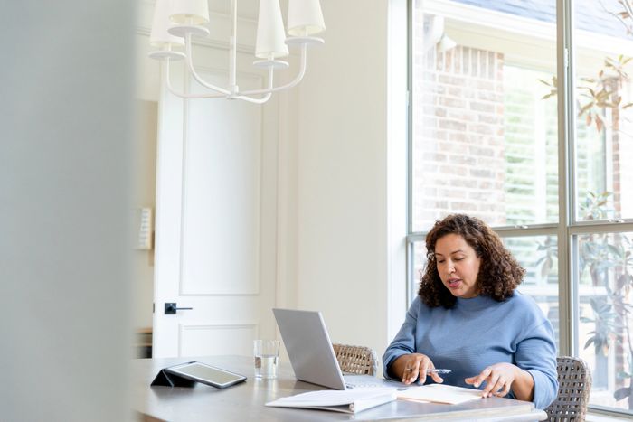 Woman working on laptop and papers at home by a window.