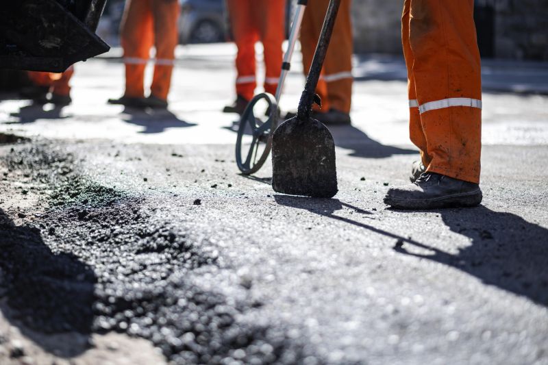 Construction crews are actively repairing a section of roadway using shovels and other tools. Bright orange work attire stands out against the asphalt. It's a clear day in a bustling urban area.