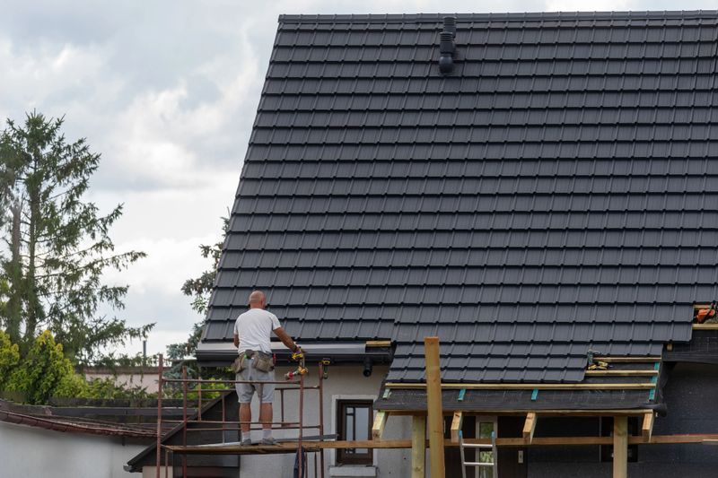 A roofer worker carefully places shingles on a sloped roof of modern house using scaffolding. Concepts of typical home renovation activity and building under construction