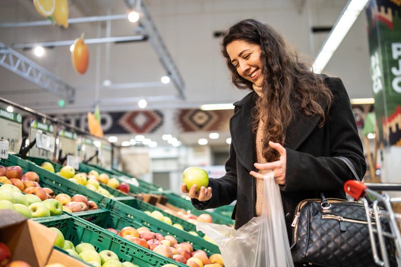 A happy woman shopping for groceries, examining a green apple at a supermarket's produce section. The vibrant fresh produce and bright interior evoke freshness, health, and sustainable living concepts.