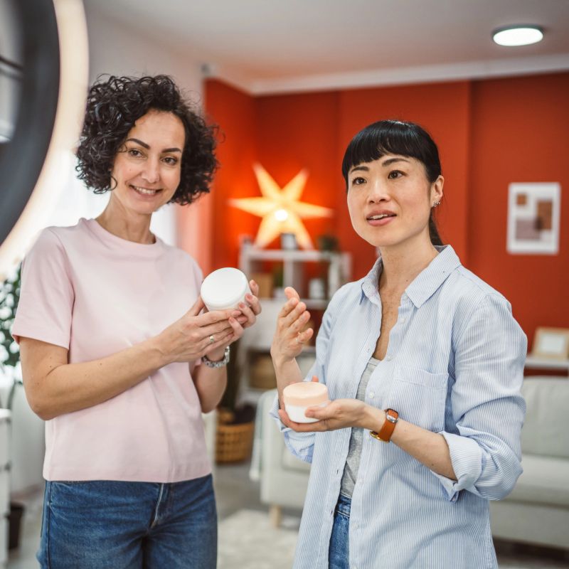 two mature multicultural woman record on mobile phone review and present skincare product for everyday use