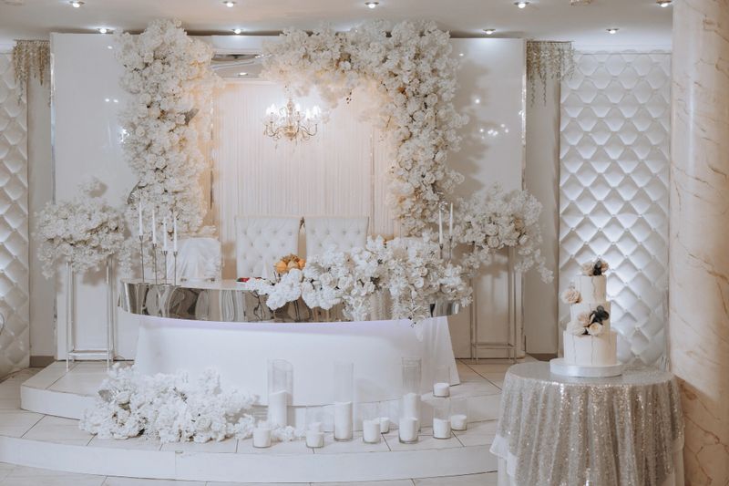 A wedding reception with a white theme. The room is decorated with white flowers and white furniture. The table is set with a cake and candles