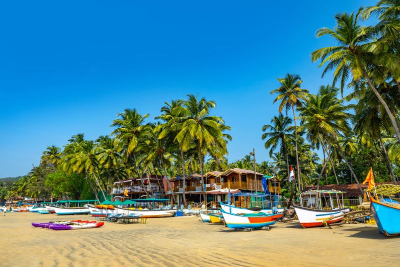 Coastal scenery featuring colorful fishing boats on a sandy beach lined with palm trees under a clear blue sky, showcasing the tropical charm of Goa, India, and evoking relaxation and serenity.