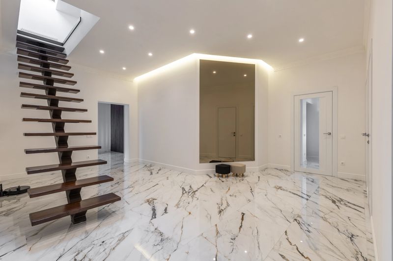 A luxurious interior featuring a floating wooden staircase, marble flooring, white walls, recessed lighting, and a large mirror, creating a sleek modern look.