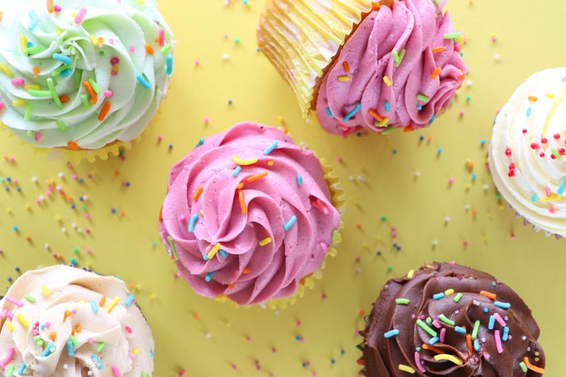 Stock photo of an overhead view of golden yellow cupcakes with smooth buttercream icing and vibrant rainbow sprinkles, with a bite taken from one. The freshly baked muffins and cakes are perfectly arranged, showcasing a delicious combination of creamy frosting and playful sprinkles