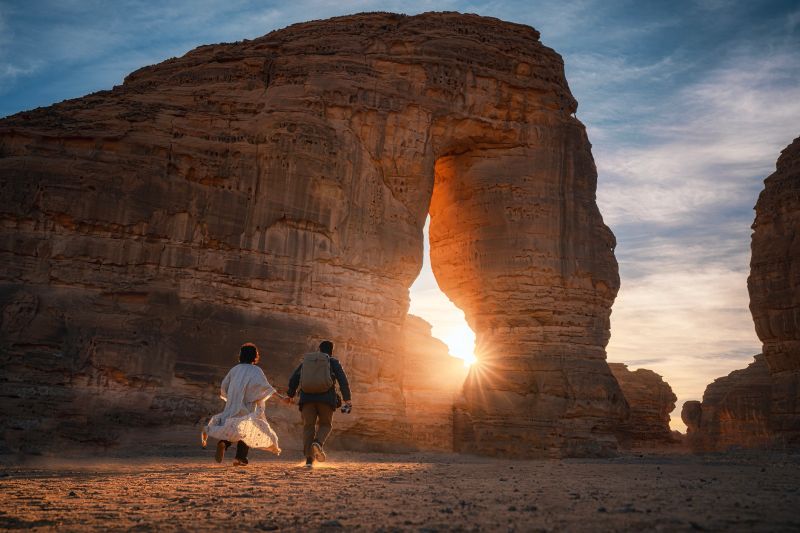 Asian Chinese couple tourist running towards Jabal AlFil - Elephant Rock in Al Ula desert landscape, dramatic sunset