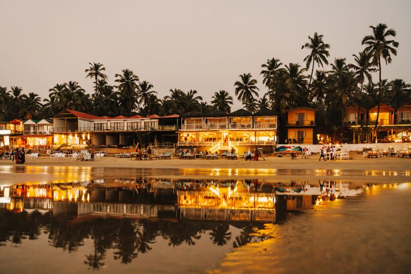 A vibrant beachfront view showcasing restaurants and serene reflections surrounded by lush palm trees during twilight in Goa, India.