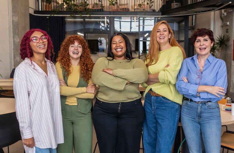 Five diverse businesswomen are standing together in their modern office, smiling confidently with their arms folded, showcasing teamwork and female empowerment