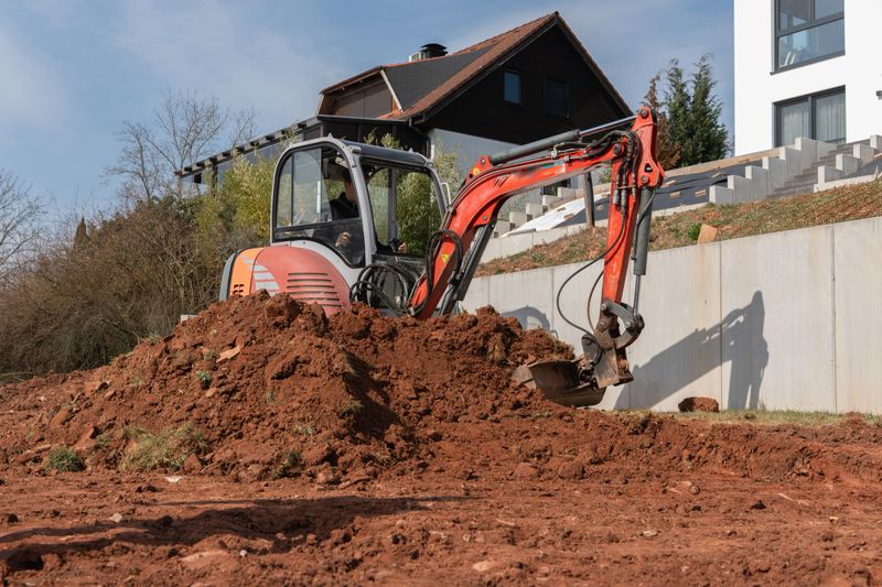 A compact excavator removes soil on a sloped plot near a modern residential building. The construction site is prepared for landscaping or foundation work. . High quality photo