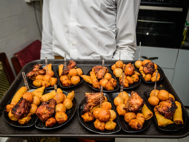 Server presenting assorted Nigerian small chops platter, featuring puff-puff, grilled chicken, samosas, and spring rolls at a social event in Lagos