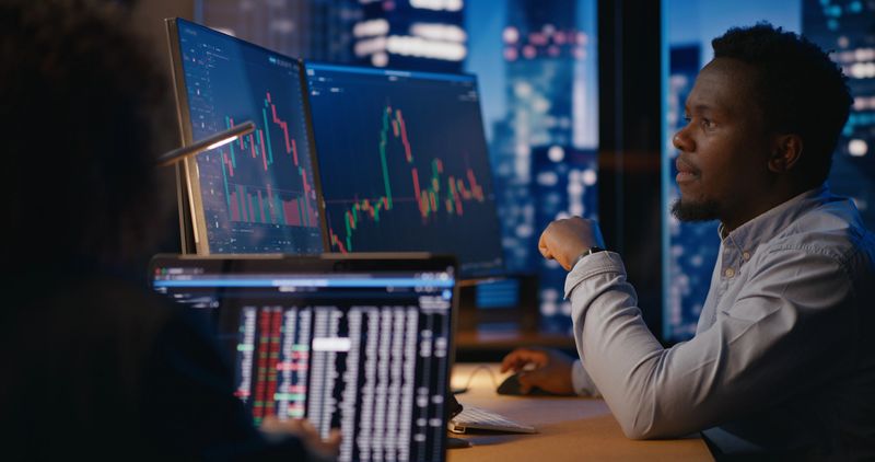 Financial advisors observe and analyze stock market charts, discussing prices, potential consequences, and developing investment strategies for the future. Modern office in skyscarper on high floor.