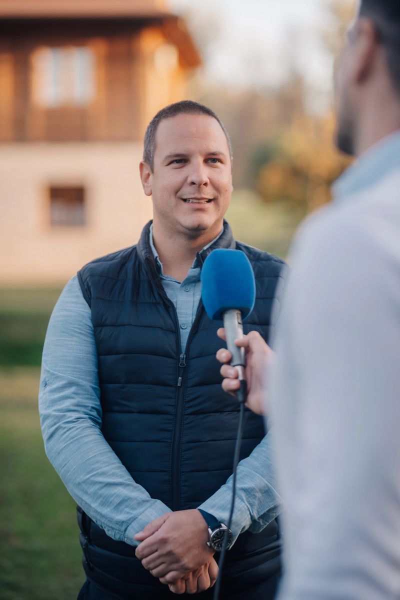 Journalist is holding a microphone and interviewing a businessman outdoors, in front of a blurred house