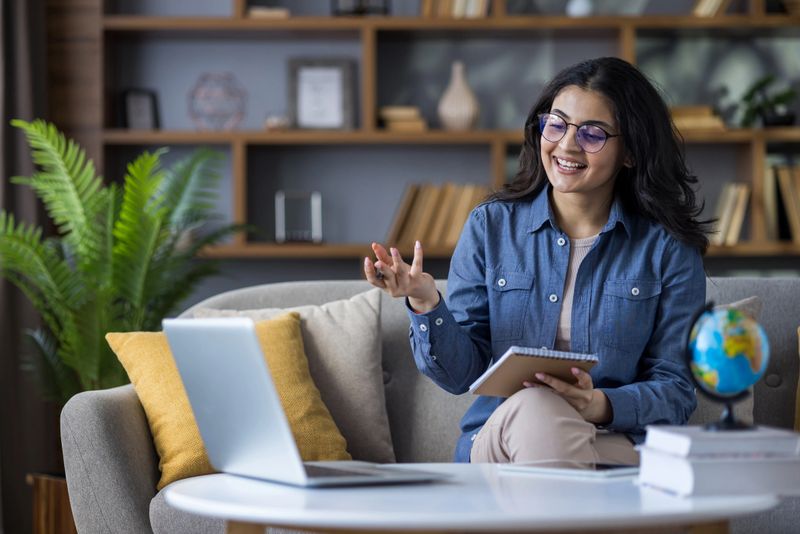 Indian young female teacher teaching remotely from home, sitting on sofa, holding notebook and books, talking via video call.