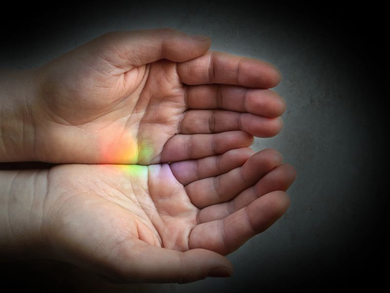 Close-up of female cupped hands with rainbow spot. Dark background, space for copy.
