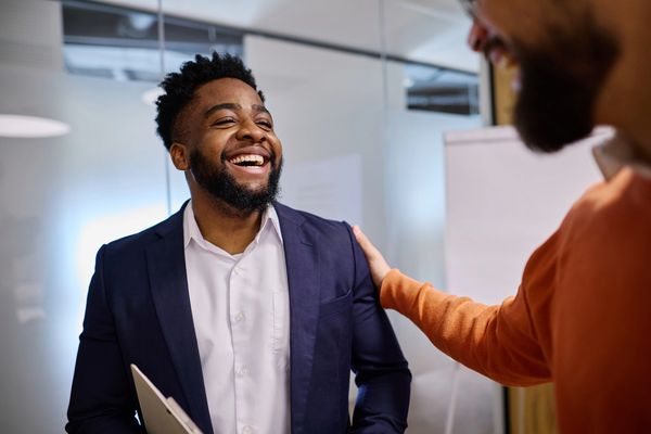 A man in a navy jacket and white shirt beign congratulated by another man in an orange jumper at work