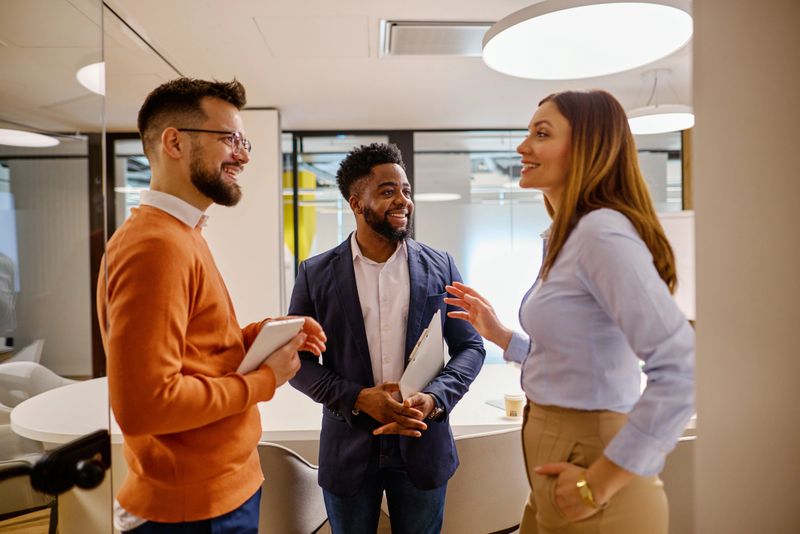 Three young businesspeople engaging in conversation in a modern office, holding tablets and documents, smiling while discussing work and collaborating on innovative ideas