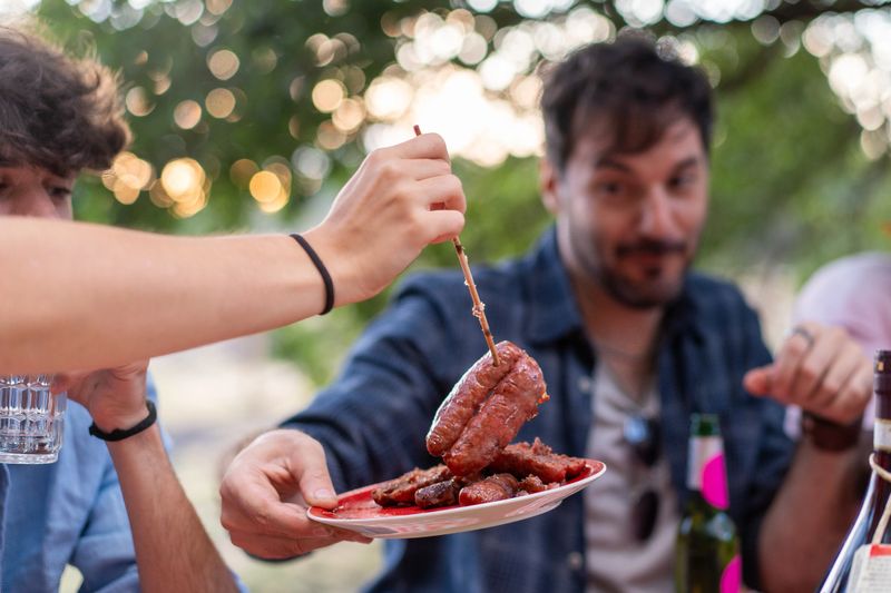 Fresh grilled sausages being shared during outdoor barbecue gathering. Natural lifestyle moment of friends enjoying summer bbq party with bokeh lights background