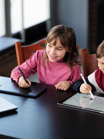 Two children happily drawing on digital tablets at a table with a laptop nearby.