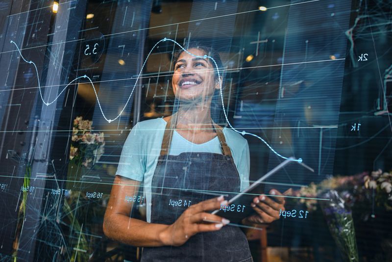 Young female florist wearing an apron and holding a digital tablet while looking at financial graphs and business analytics overlaid on the store window, symbolizing small business growth, technology integration, and entrepreneurship.