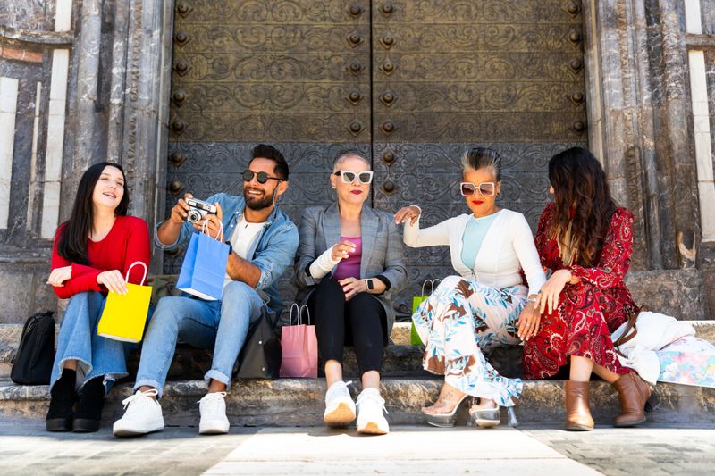 Happy multi-ethnic group of friends resting on steps after a shopping spree, taking pictures and smiling