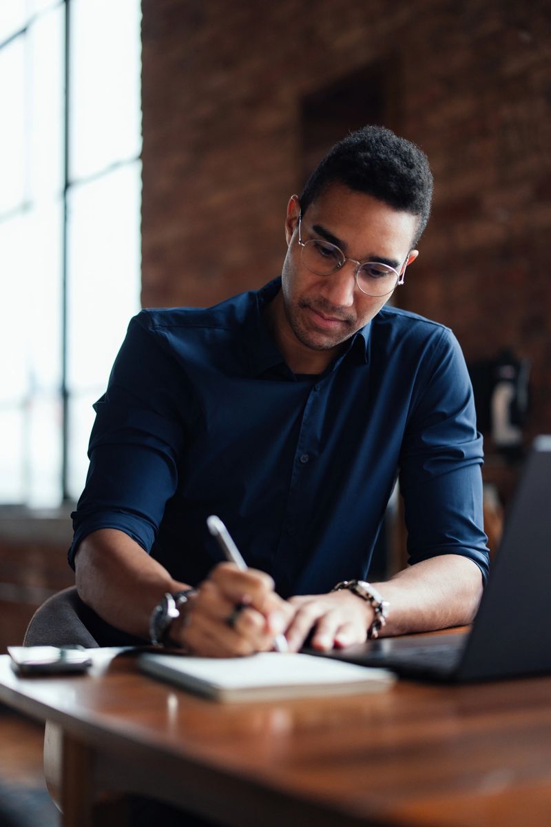 A professional seated at a desk in a well-lit, modern office space, engrossed in writing notes with a considered expression, embodying productivity and focus within a contemporary co-working environment.