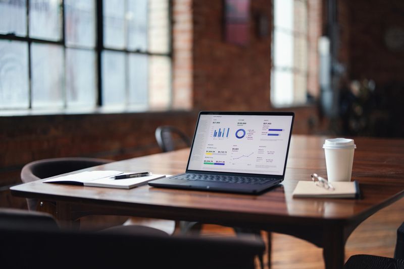 Laptop showcasing detailed data analytics placed on a rustic desk in a contemporary office environment with natural light from large windows, emphasizing productivity, technology, and professional working atmosphere.