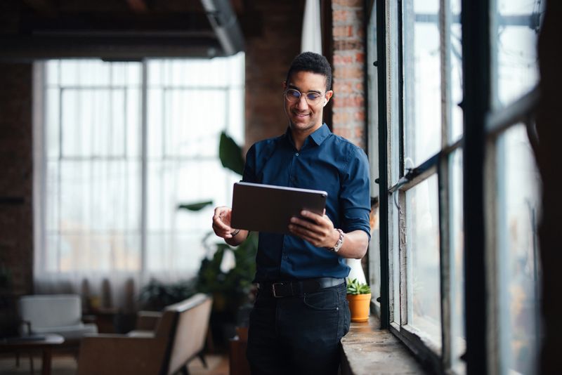 A professional man stands near a window in a modern and stylish workspace, engaged with his tablet. The scene depicts a productive atmosphere with cozy furnishings and a natural light-filled environment.