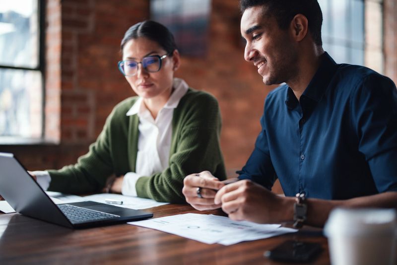 Two colleagues collaborate on a project in a stylish office environment with warm sunlight and brick walls. They are working together with documents and a laptop, emphasizing teamwork and productivity.