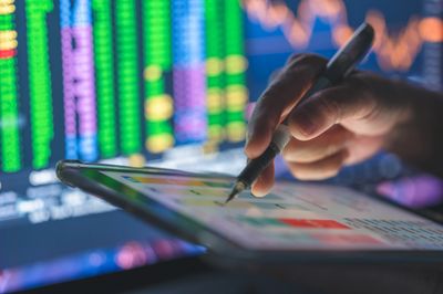 Person analyzing financial data on a digital tablet with stock market charts in the background.