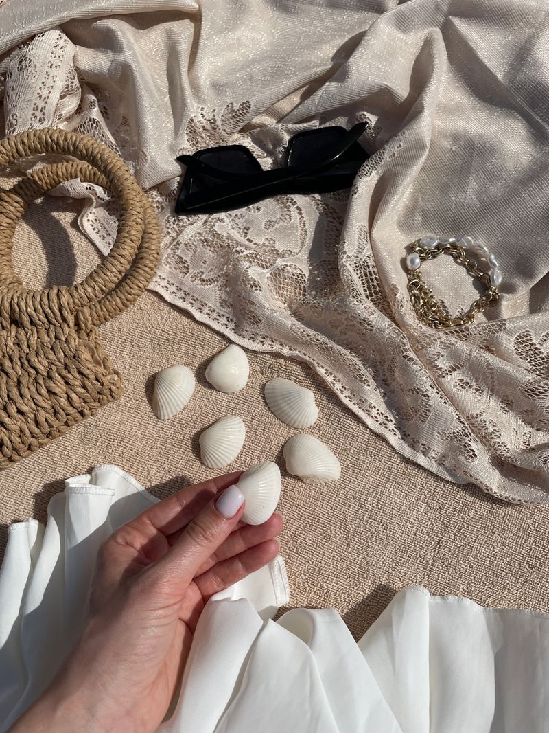 Hand holding a white seashell on a towel with beach accessories, sunglasses, wicker bag, lace fabric, and white dress