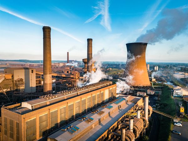 Industrial factory with smokestacks and cooling tower emitting steam under a blue sky.