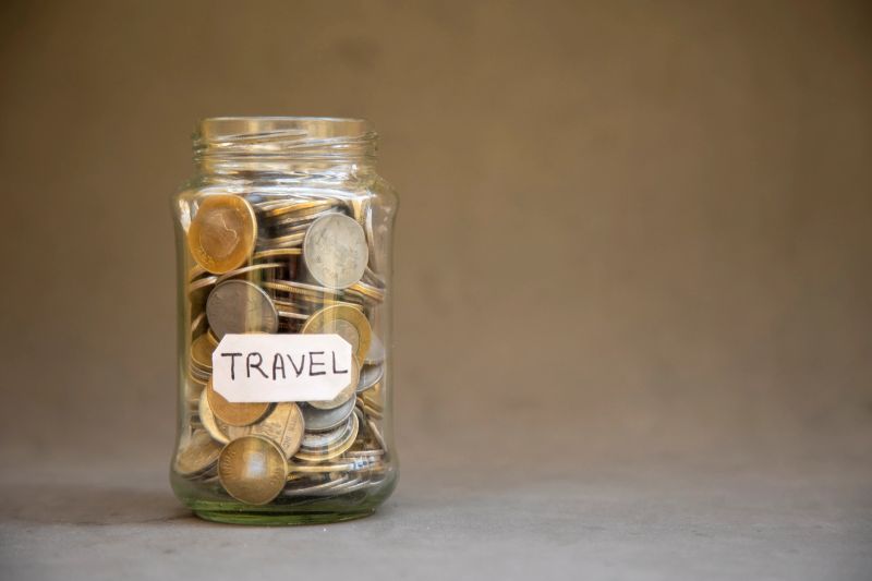 A jar holding coins labeled 'travel,' representing saving money for vacation purposes against a simple background.