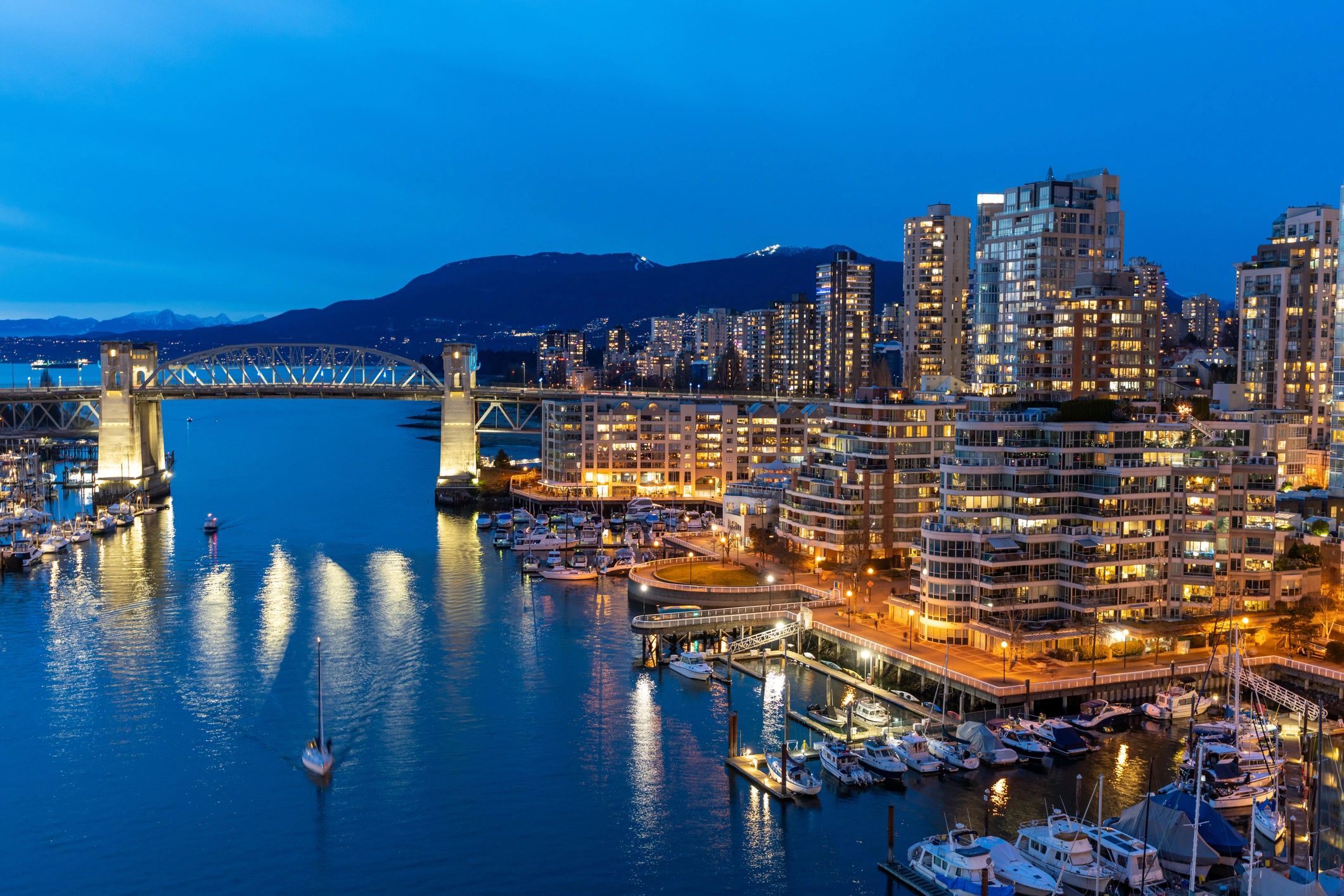 Night view of a city harbor skyline with lit high-rise buildings, illuminated bridges, and boat refl